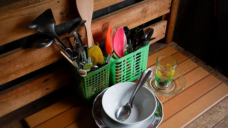 Peralatan makan dan masak rumah tangga di keranjang Sendok, garpu, sutil, centong, dan spatula berdiri di dua keranjang plastik hijau; di sebelahnya mangkuk, piring, dan gelas kaca di atas meja kayu
