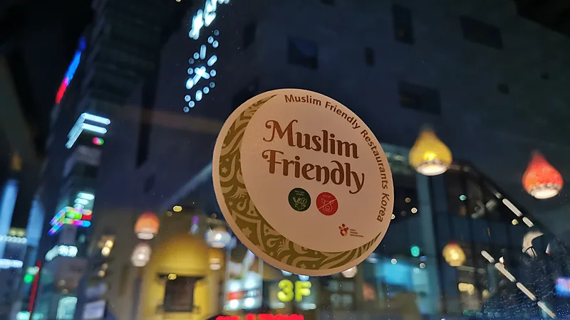 Stiker Muslim Friendly di kaca restoran Stiker bundar krem bertulisan Muslim Friendly Restaurants Korea menempel di kaca sebuah restoran, dengan lampu kota Korea Selatan menyala di belakangnya