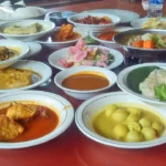 Belasan piring lauk masakan Padang tersaji di meja merah: gulai ayam, rendang, ikan goreng, dan telur berkuah kuning
