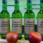 Empat botol kaca hijau cuka apel organik berlabel kertas cokelat berdiri di peti kayu, dengan dua apel merah di depannya